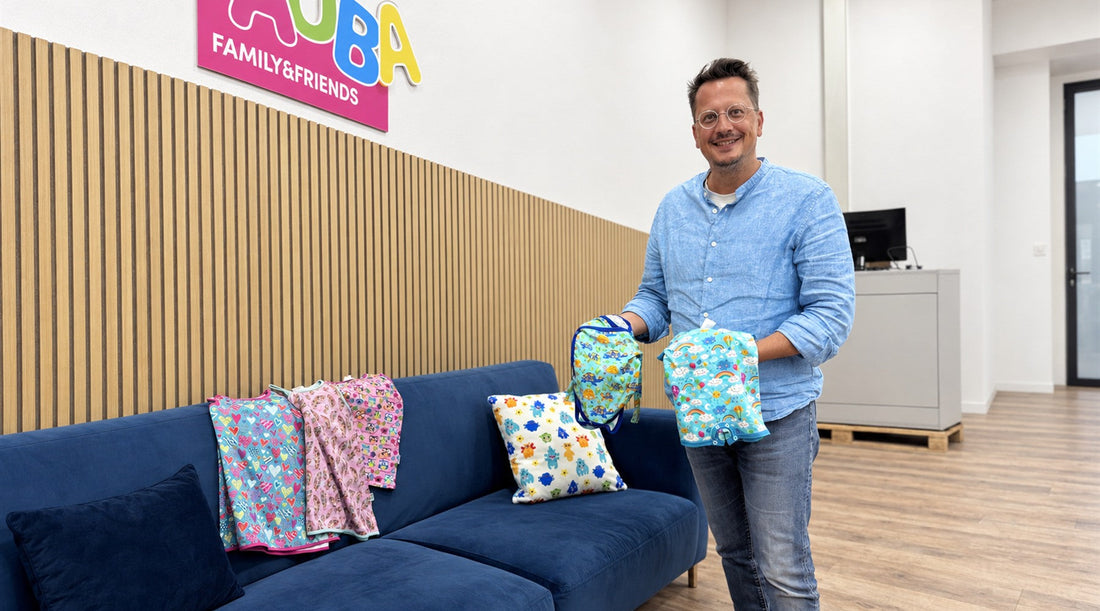 Lächelnder Mann präsentiert farbenfrohe, handgefertigte Kinderprodukte vor einem blauen Sofa.