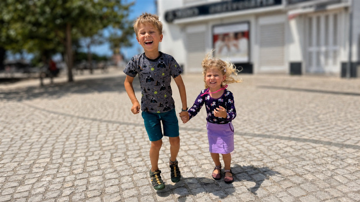 Zwei lachende Kinder springen Hand in Hand in gemusterter Babauba-Kinderkleidung.