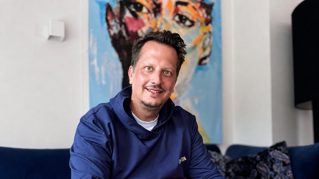 Patrick Wallinger sitzt lächelnd in einem dunkelblauen Hoodie auf einem blauen Sofa vor einem farbenfrohen Gemälde.